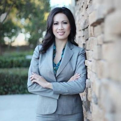 Woman in gray suit, arms crossed, leaning against a stone wall, smiling outdoors.
