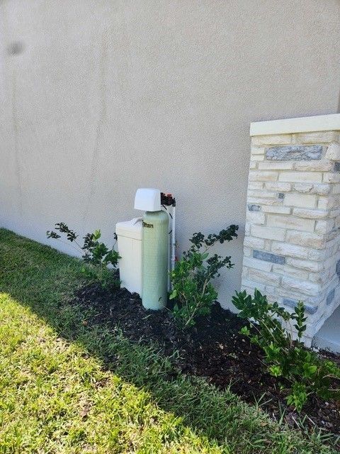 Green water softener unit next to a light-colored stucco wall and a stone-covered column, with landscaping.