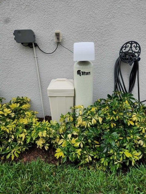 Water softener and tank next to beige utility box against a stucco wall, surrounded by green and yellow shrubs.