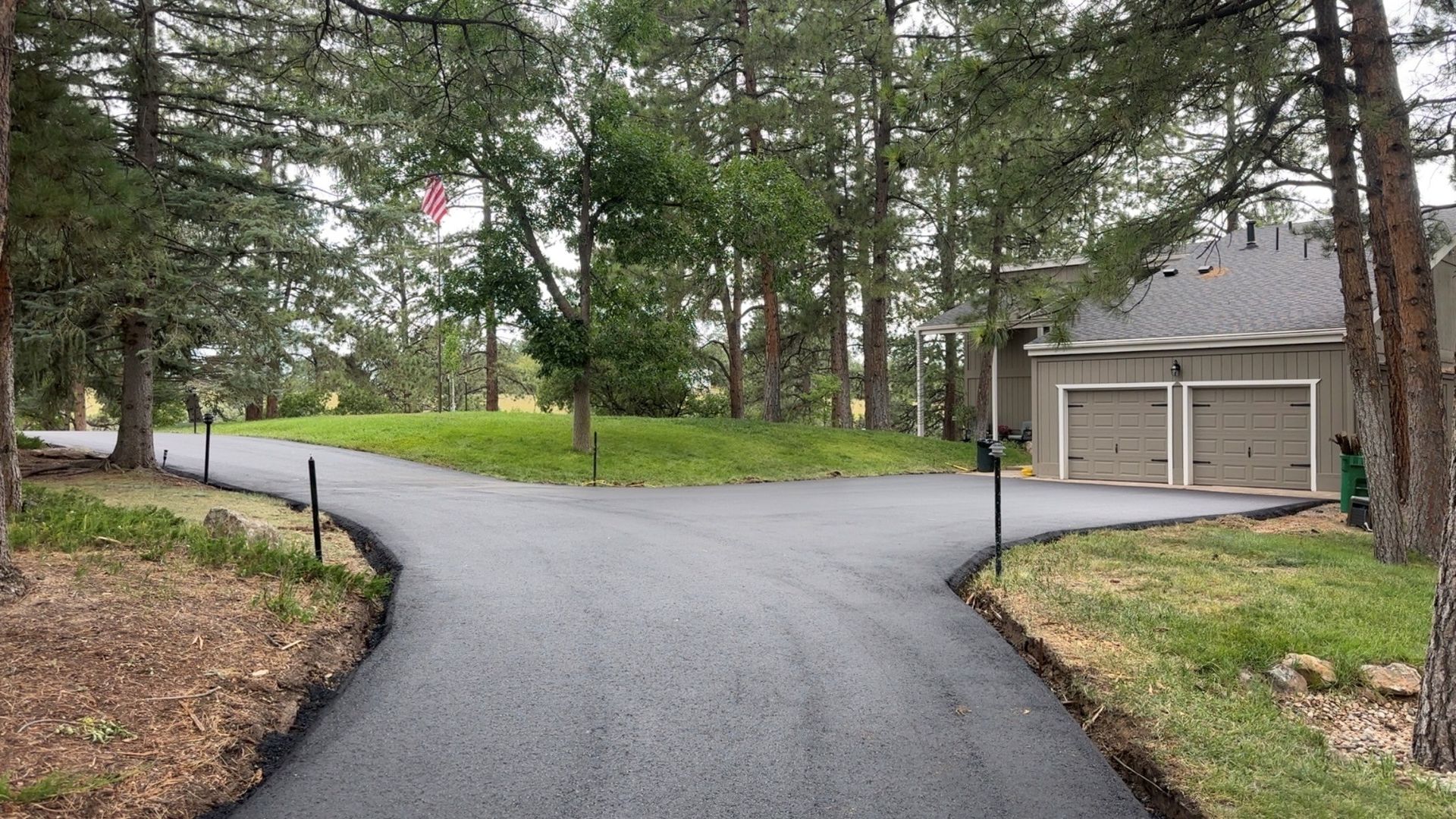 Paved driveway forks, leading to a garage and a grassy hill, surrounded by trees.