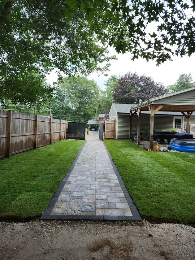 A stone paver walkway with a dark border runs between green lawns, leading to a wooden fence and a covered patio area.