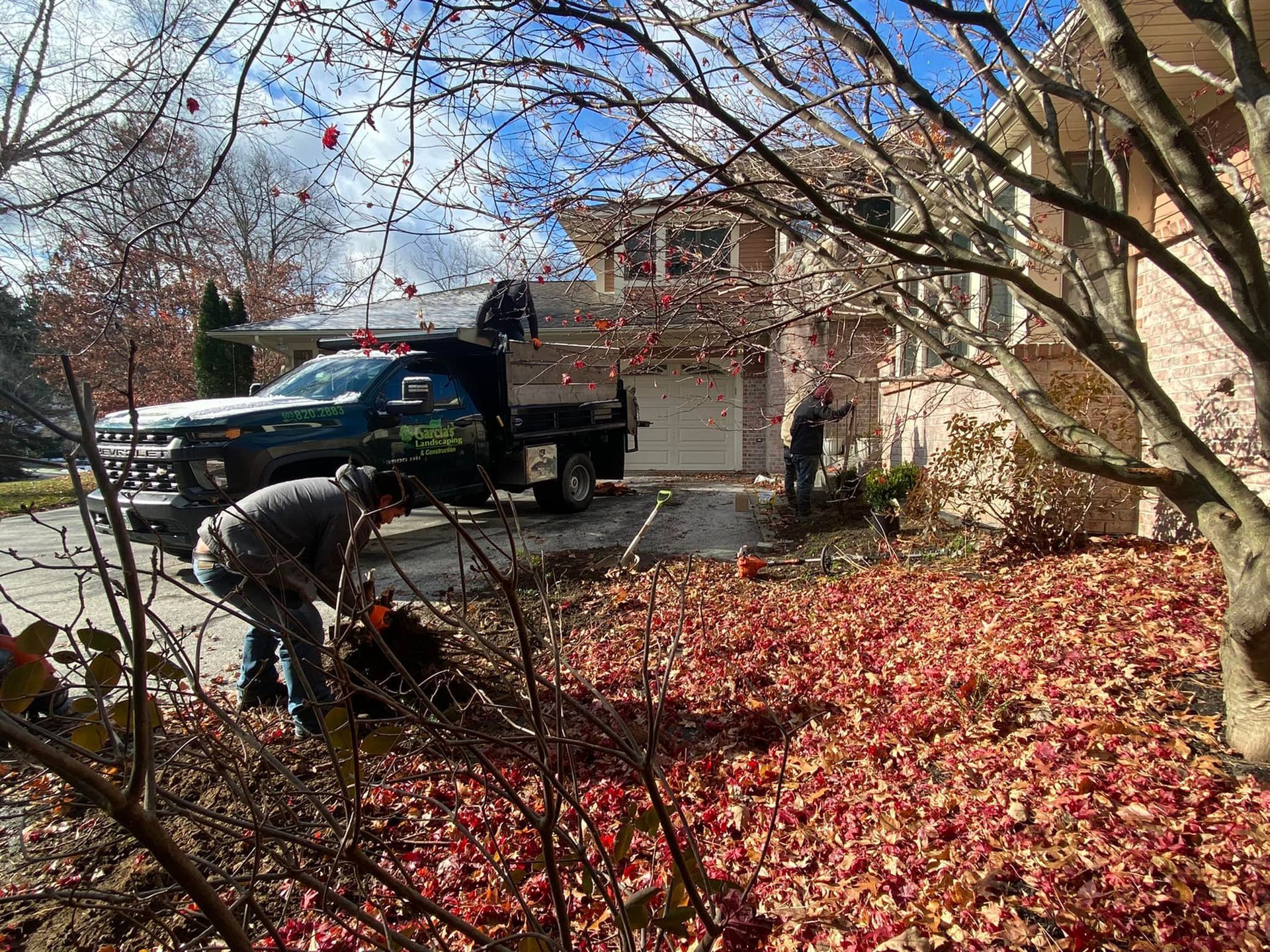 Two workers clear fallen red leaves from a residential yard with a dump truck parked in the driveway.