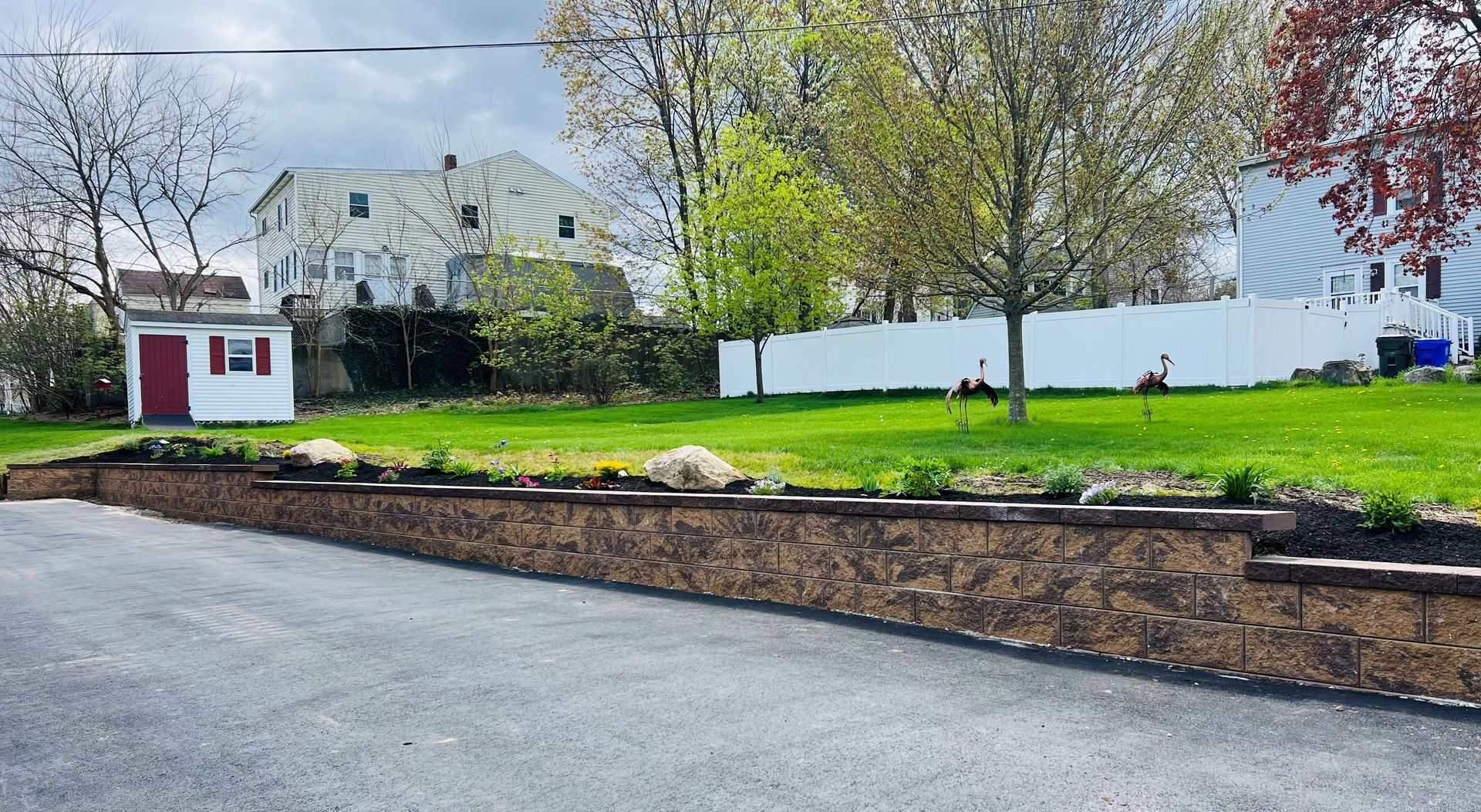 A low stone retaining wall borders a paved driveway in front of a green lawn, a white fence, and houses.