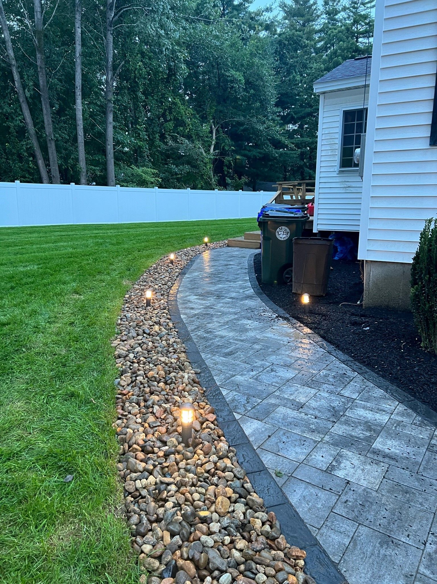 A stone paver walkway illuminated by small lights, bordered by decorative rocks and a green lawn next to a white house.
