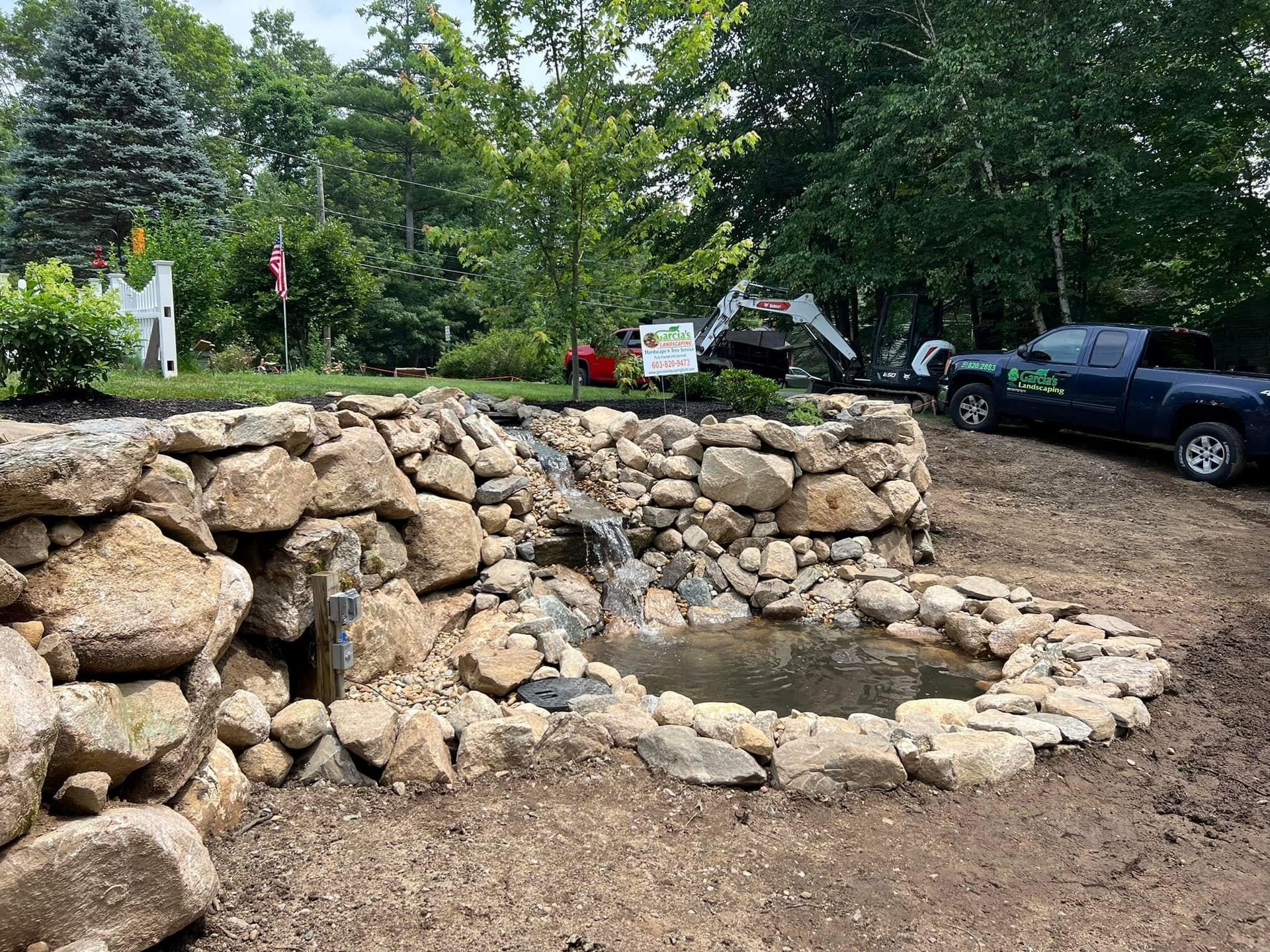 A natural stone waterfall feature flows into a small basin, set in a woodsy landscape with a parked truck in the background.