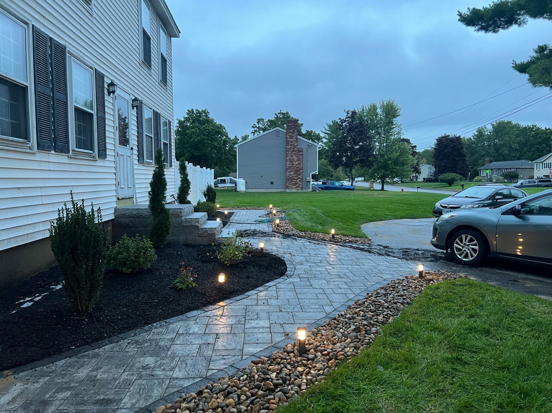 A paved stone walkway illuminated by low-voltage lights leads to a suburban house with white siding at dusk.