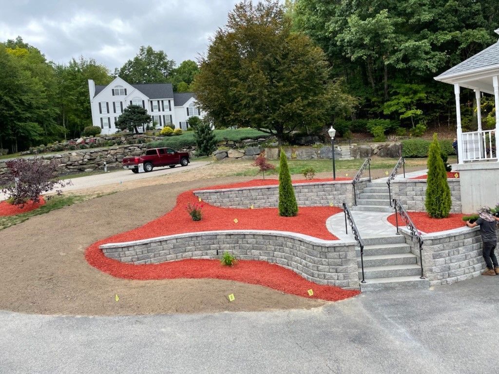 Stone retaining walls with red mulch and new landscaping stairs lead to a white house with a red truck parked in the drive.