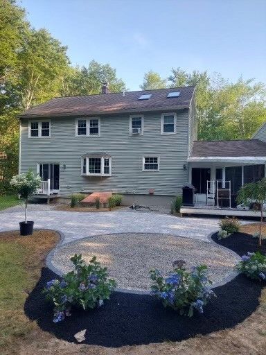 A two-story light-gray house with a paved circular patio, black mulch beds with blue hydrangeas, and a gravel yard.