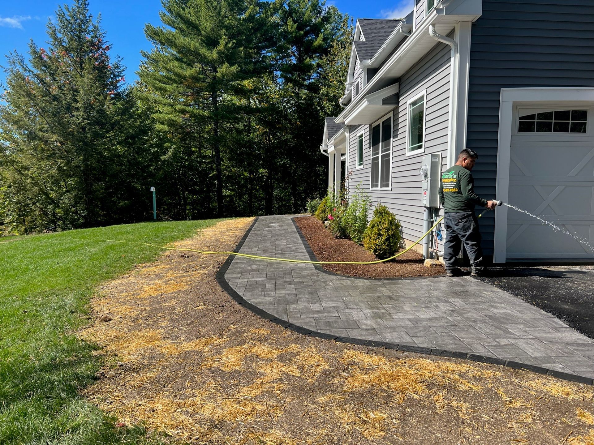 A worker sprays a newly installed paver walkway next to a gray house and a grass lawn under a bright blue sky.