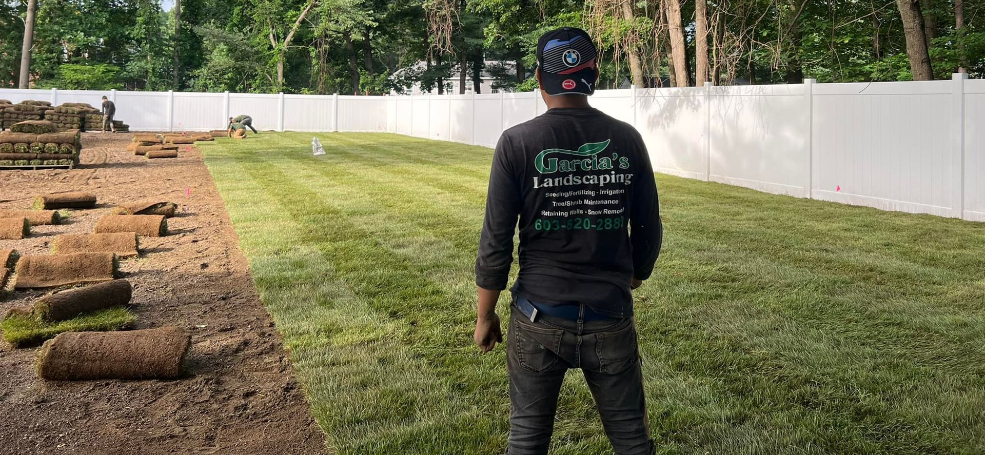 A landscaper stands in a yard after installing new sod rolls next to a freshly laid green lawn and white vinyl fence.