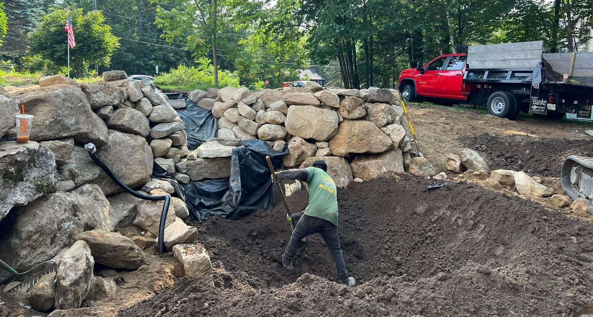 A worker in a green shirt digs into the soil in front of a newly constructed stone retaining wall near a red truck.