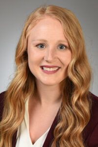 Woman with long, wavy auburn hair smiles at the camera, wearing a dark red blazer over a white blouse.