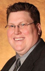 Man smiling, wearing glasses, a suit, and a tie.