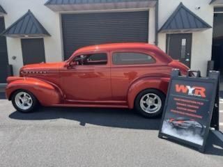 Rust-colored vintage car parked outside a building, next to a sign for WYR Auto, a paint and body shop.