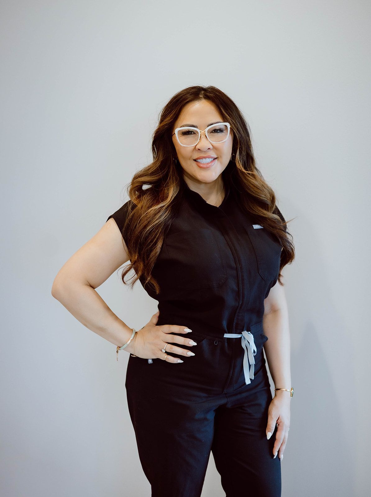 A woman wearing glasses and a black scrub suit is standing with her hands on her hips.