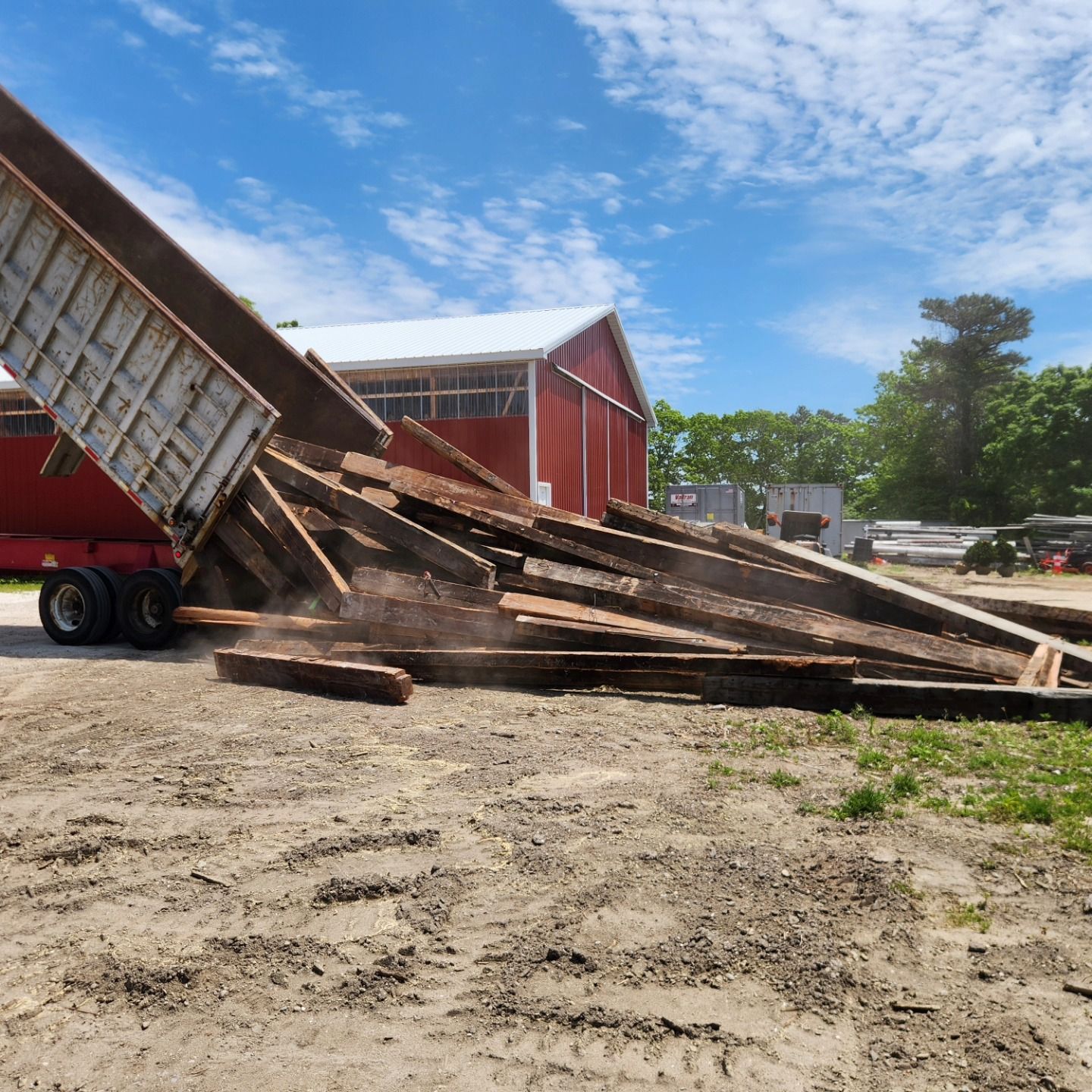 A dump truck is loading wood into a pile in front of a barn