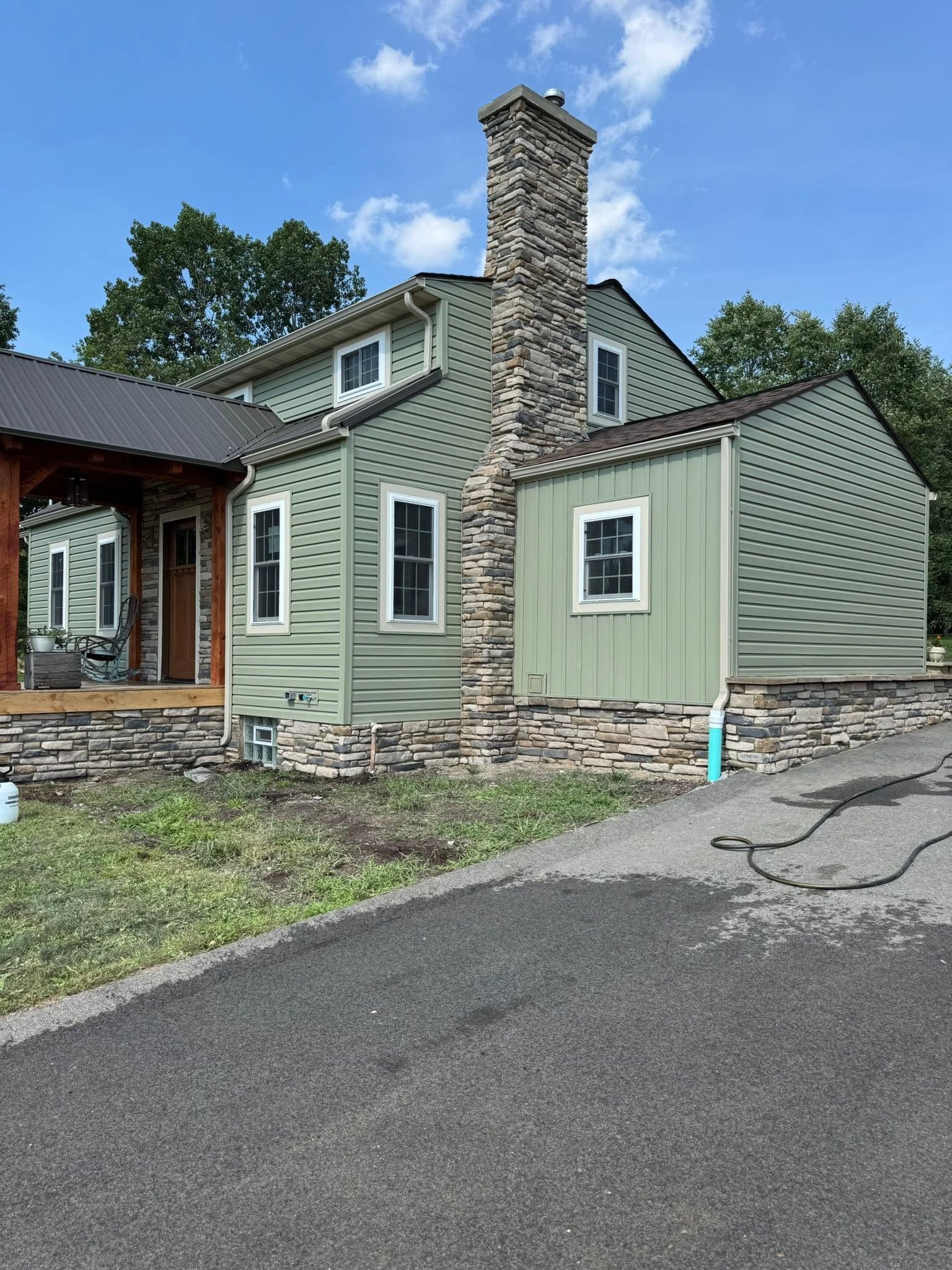 Small green house with stone chimney, white-trimmed windows, and a curved side wall beside a paved driveway.