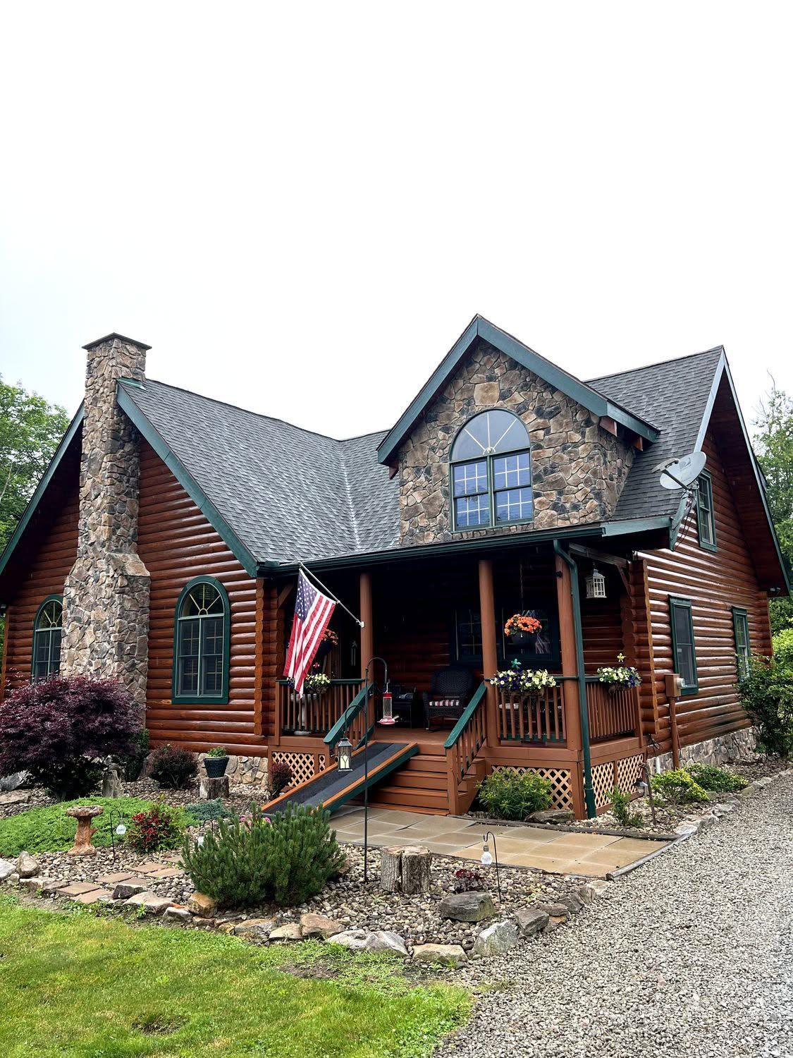 Rustic log cabin with stone chimney, covered porch, and American flag, set beside a gravel drive.