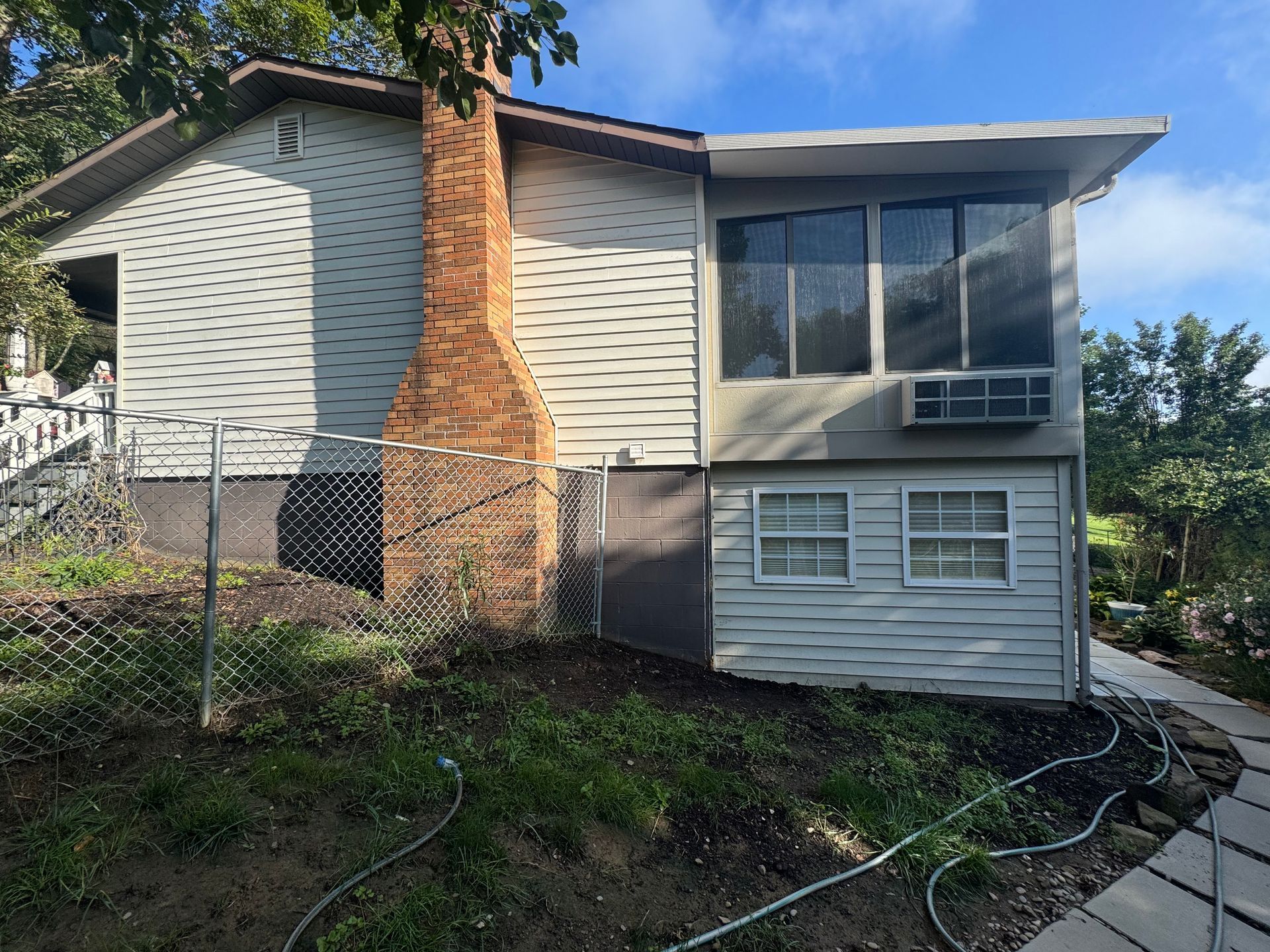 Exterior of a beige house with a brick chimney, chain-link fence, and window air conditioner on a sloped yard