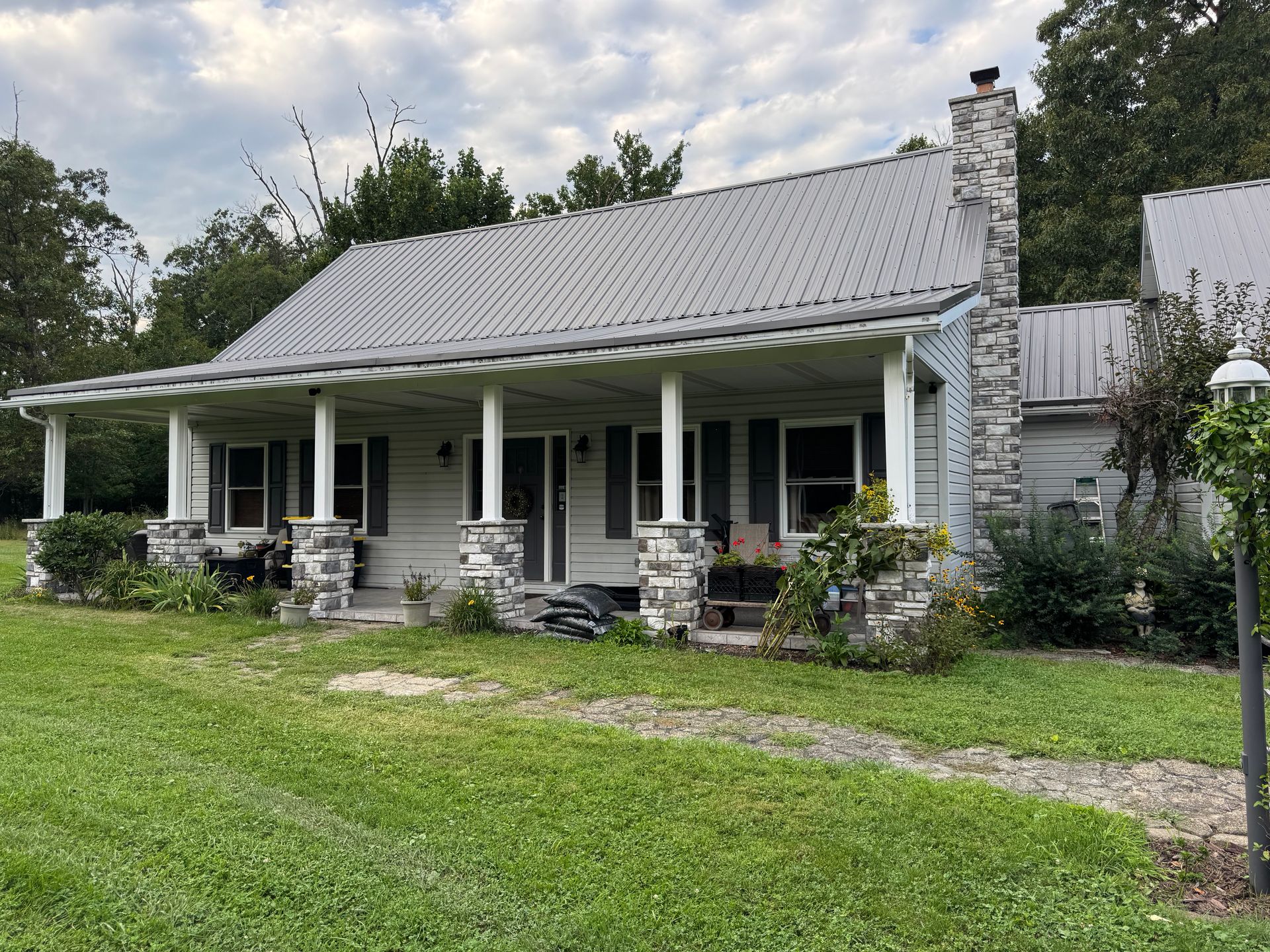 White farmhouse with a porch, stone chimney, and rocking chairs, set in a green yard under cloudy skies