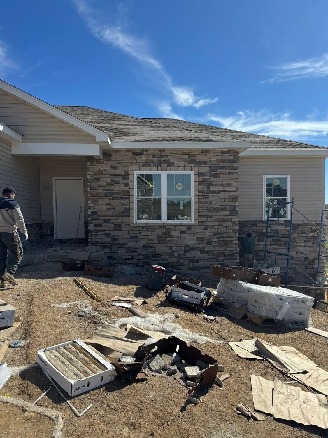 Front of a house under construction with debris scattered across the dirt yard under a blue sky