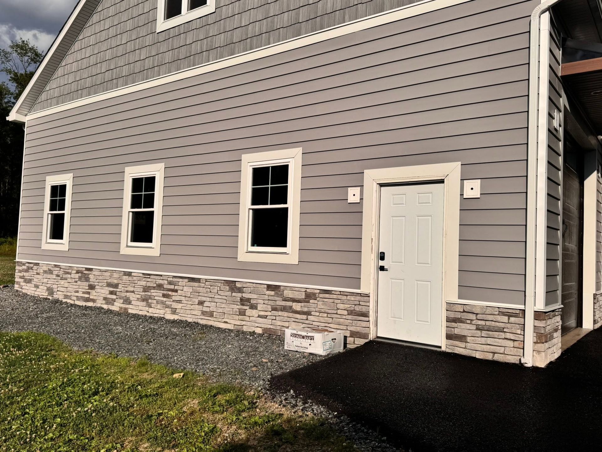 Gray house exterior with white-trimmed windows, stone base, and a white door beside a black driveway