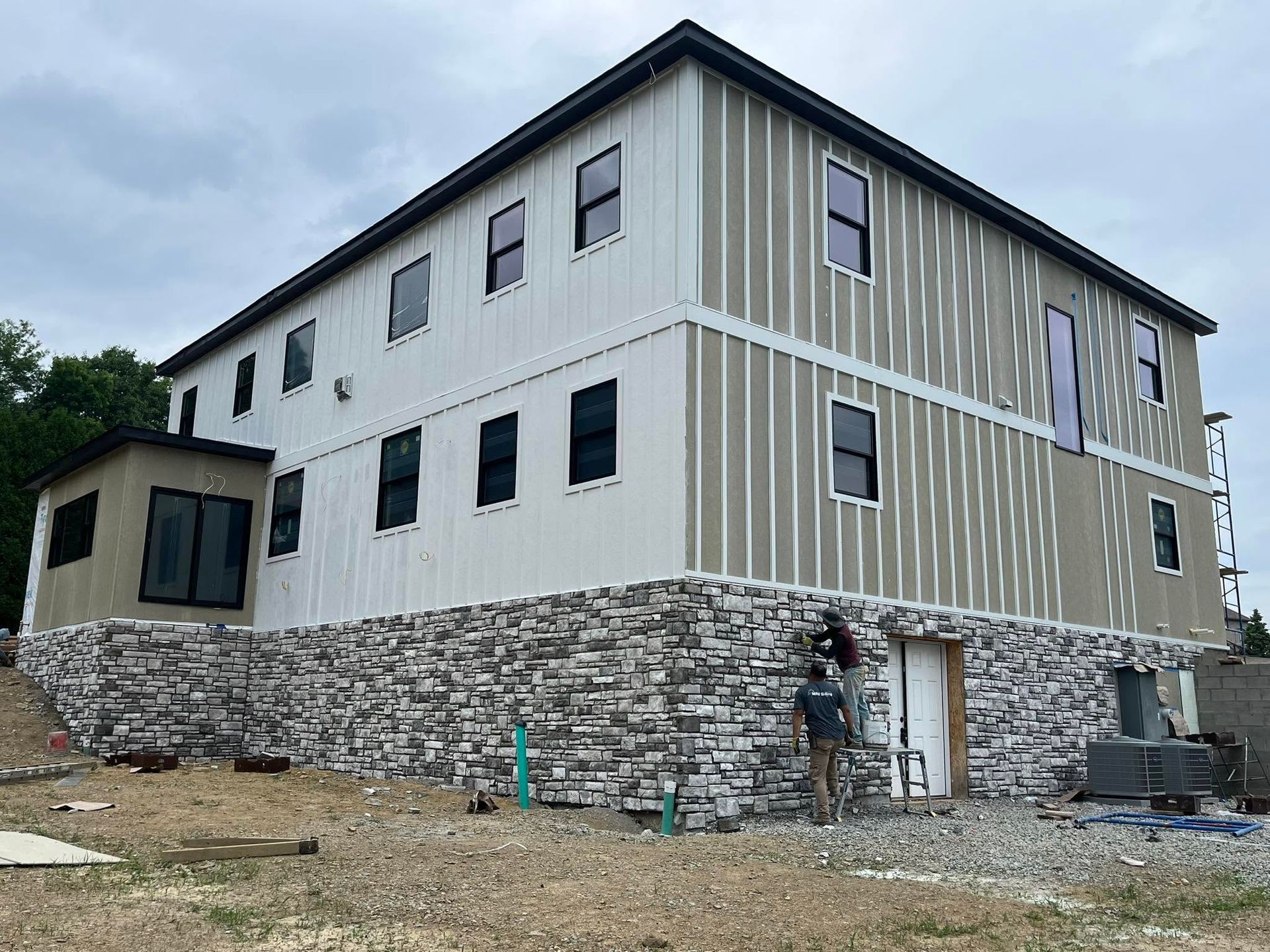 Two-story house under construction with stone foundation and gray siding under a cloudy sky