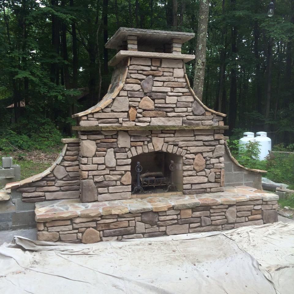 Stone outdoor fireplace built into a landscaped wooded area, with a paved foreground and forest behind