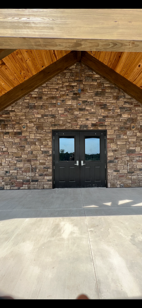 Stone entryway with black double doors, wood beams, and a concrete porch under a peaked roof.