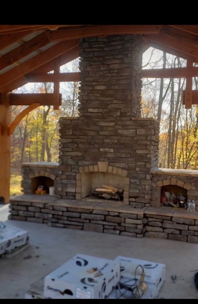 Rustic stone fireplace with three fireboxes under a wooden roof, in an open pavilion with large windows.