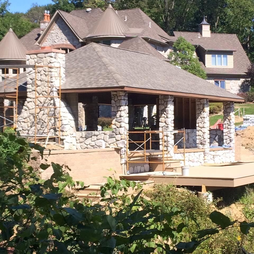 Stone gazebo under construction in a wooded residential area with houses in the background