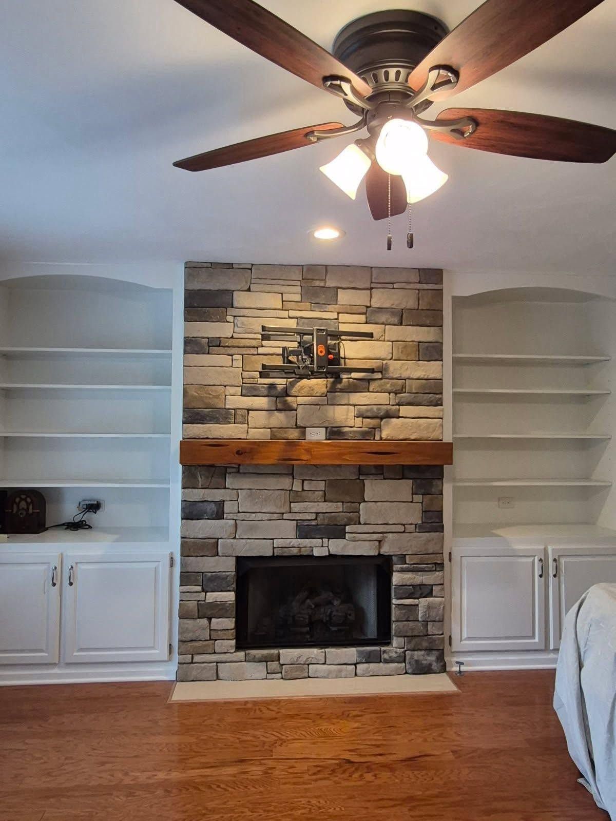 Living room with stone fireplace, white built-in shelves, wood floor, and ceiling fan above