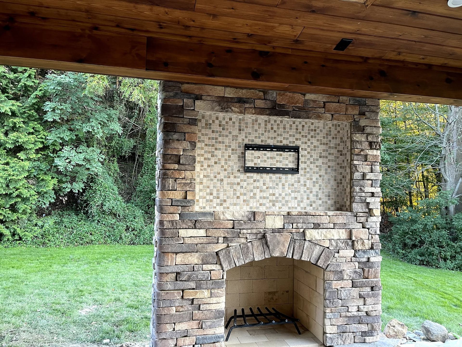 Stone outdoor fireplace under a wooden roof, set in a grassy yard with trees behind it