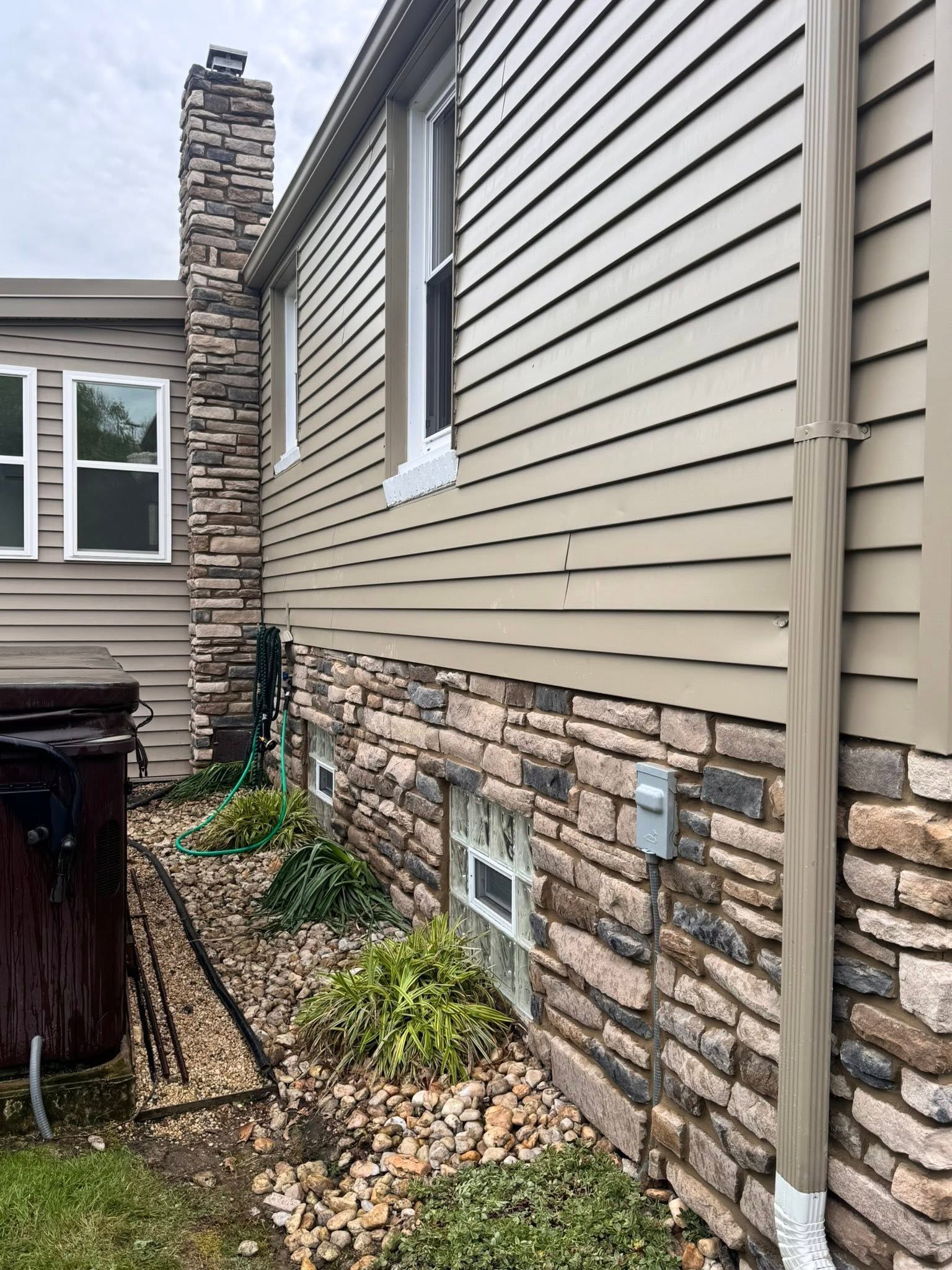 Side of a beige house with stone foundation, window, downspout, and a small landscaped bed along the wall