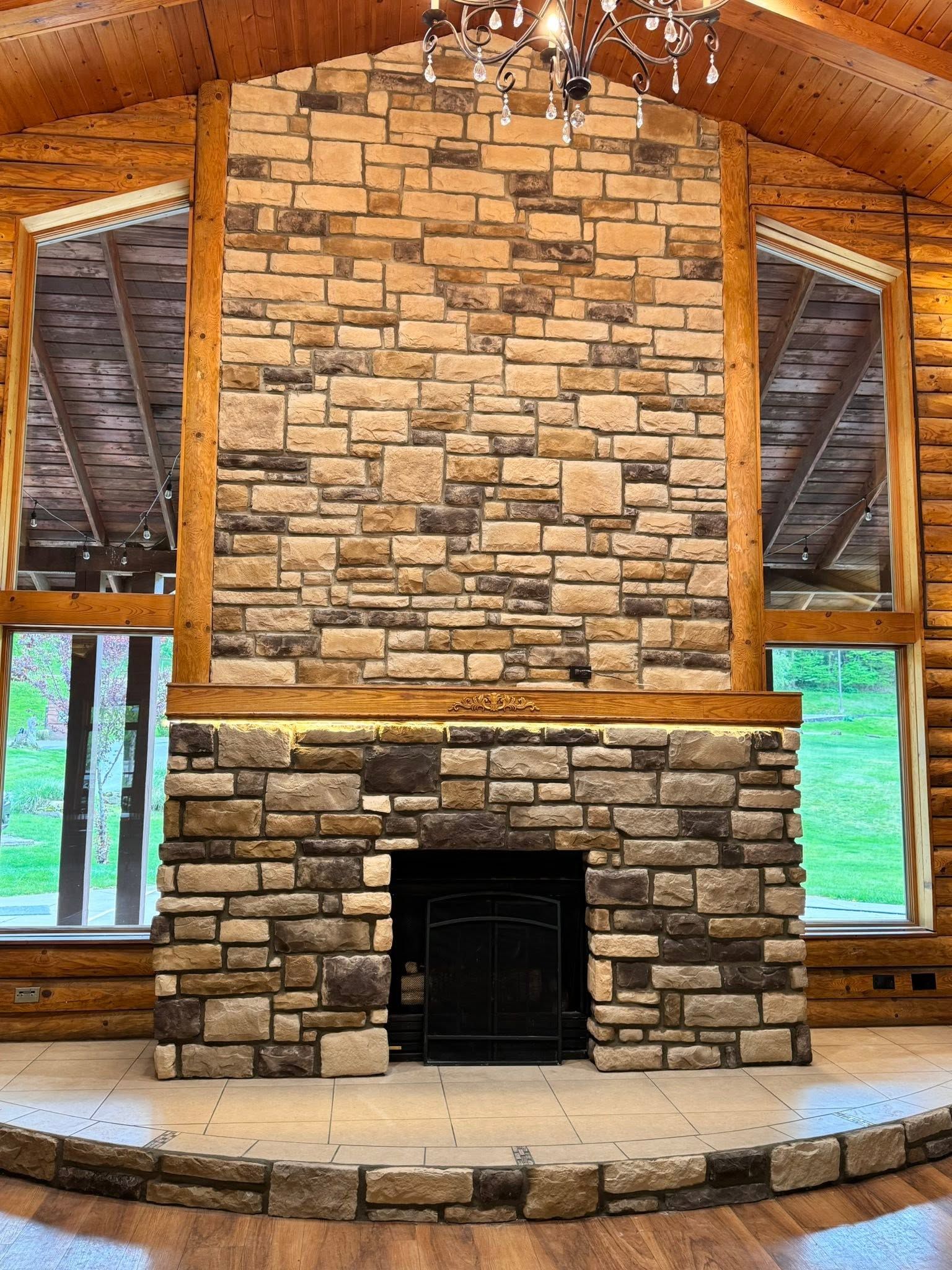 Stone fireplace centered in a rustic wooden cabin with large windows and exposed timber ceiling.