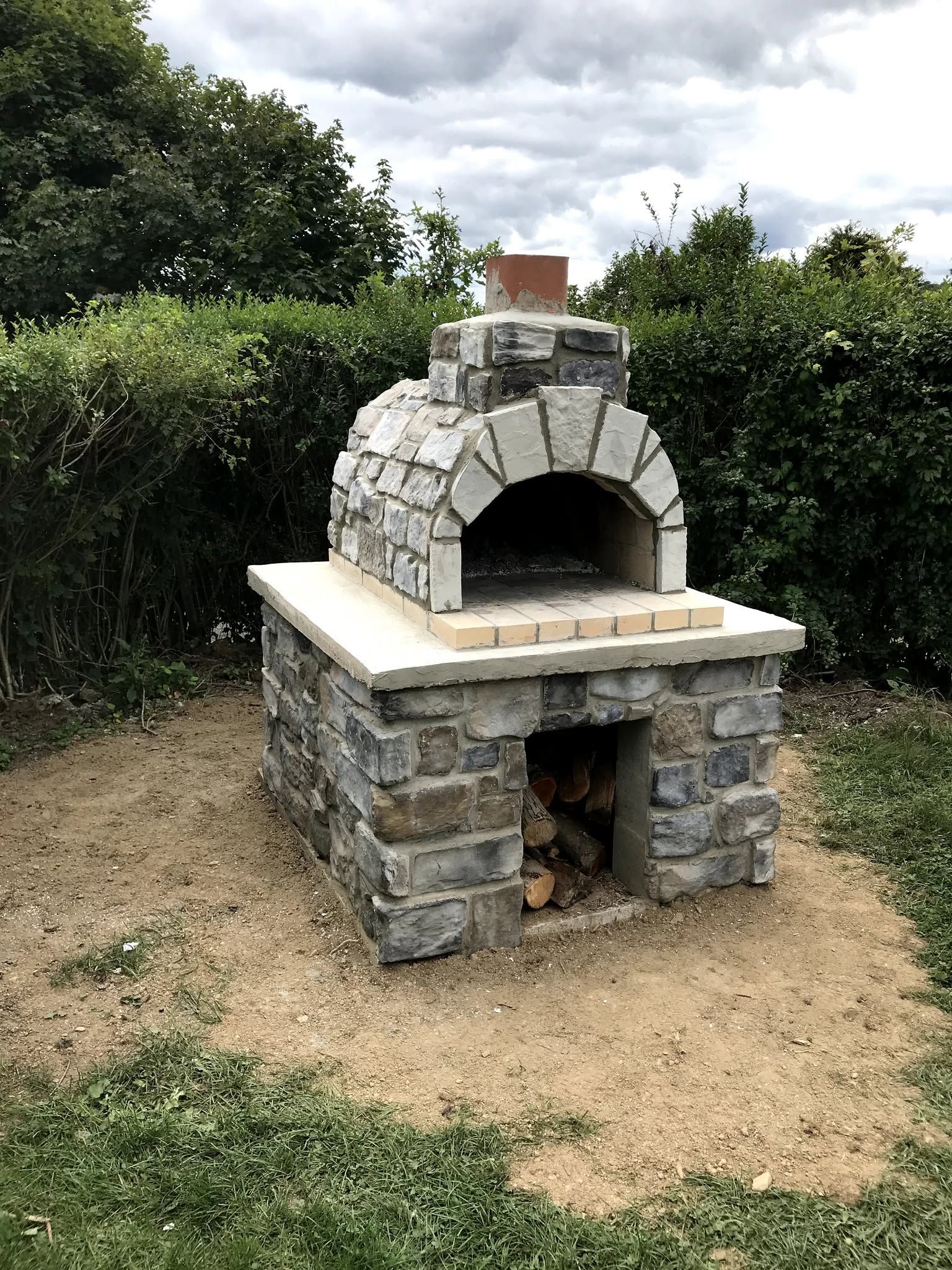 Outdoor stone brick oven with arched opening in a garden, surrounded by hedges and grass