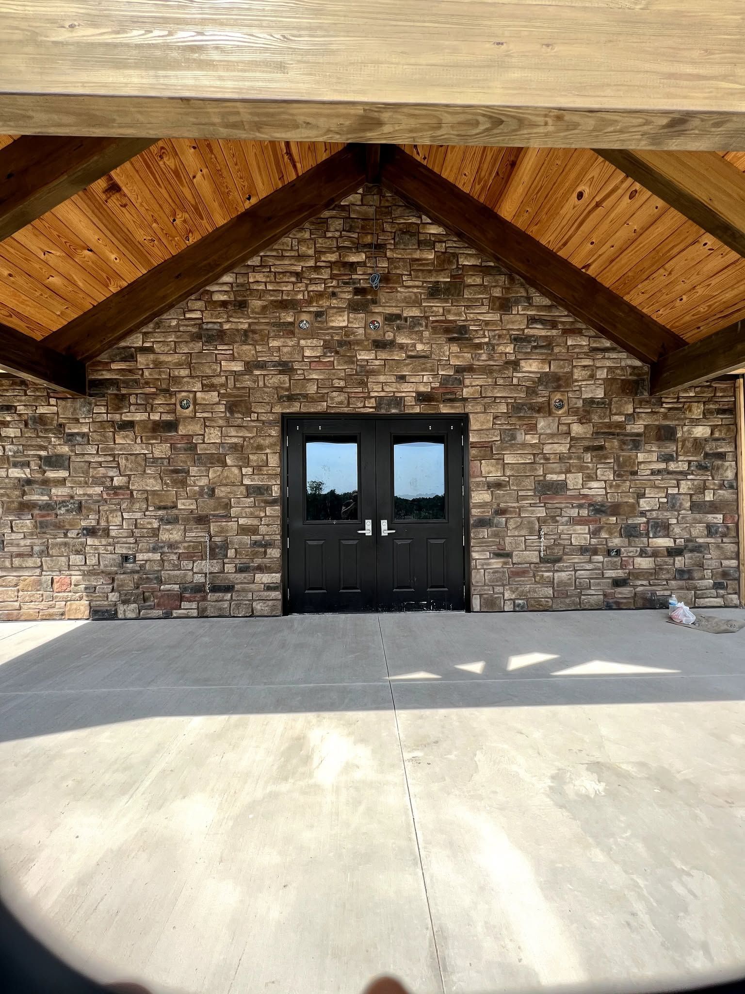 Stone building entrance with black double doors beneath a wooden gabled awning and concrete patio