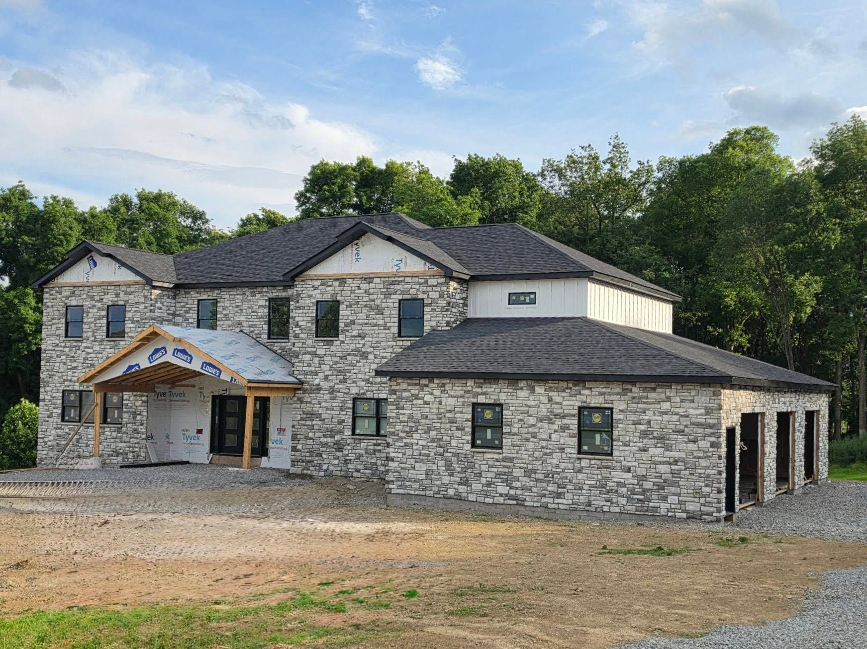 Stone house under construction with dark shingle roof and a covered front entry, surrounded by bare ground and trees