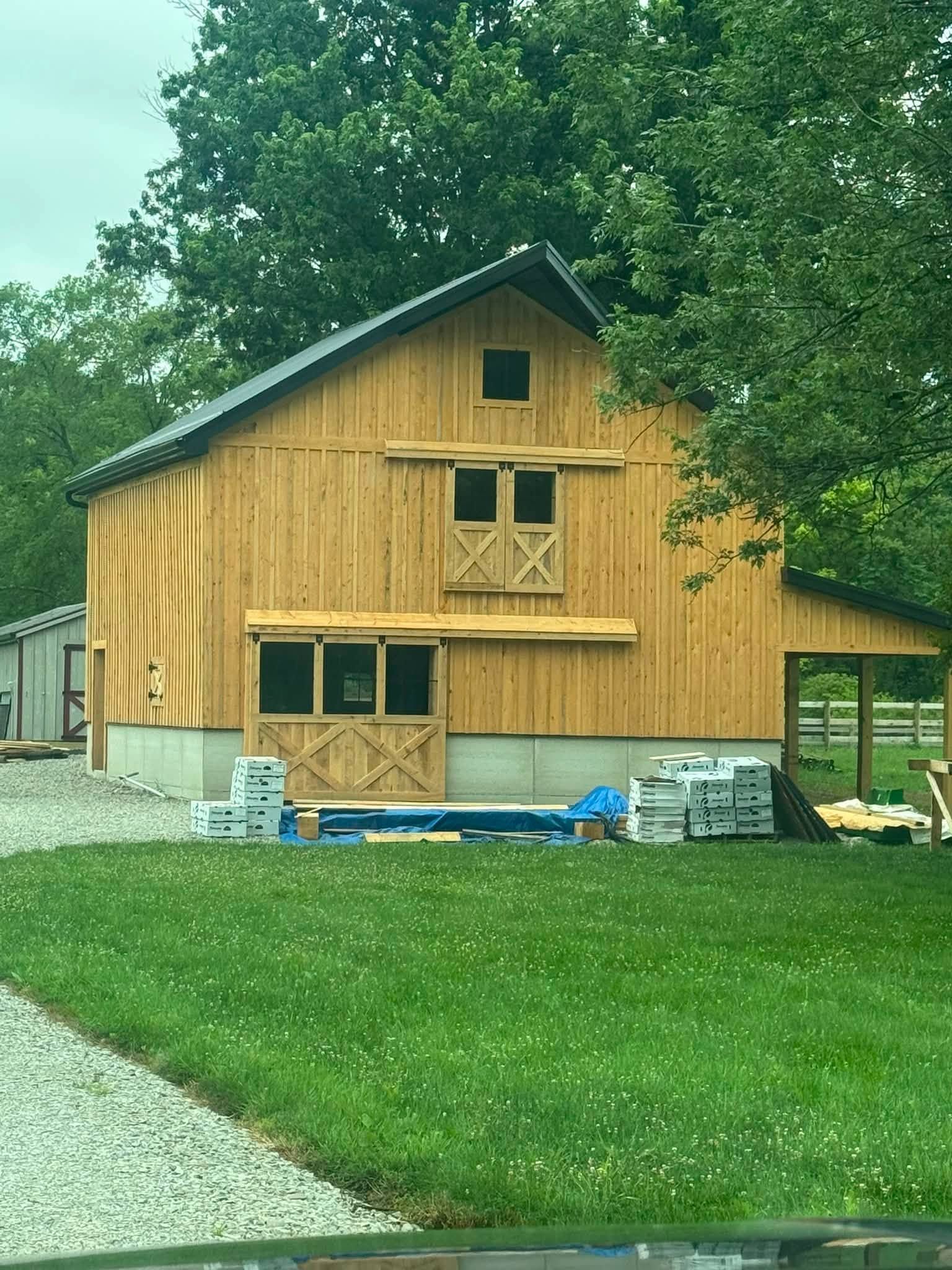 Yellow wooden barn under construction beside a green lawn and gravel drive, with trees in the background