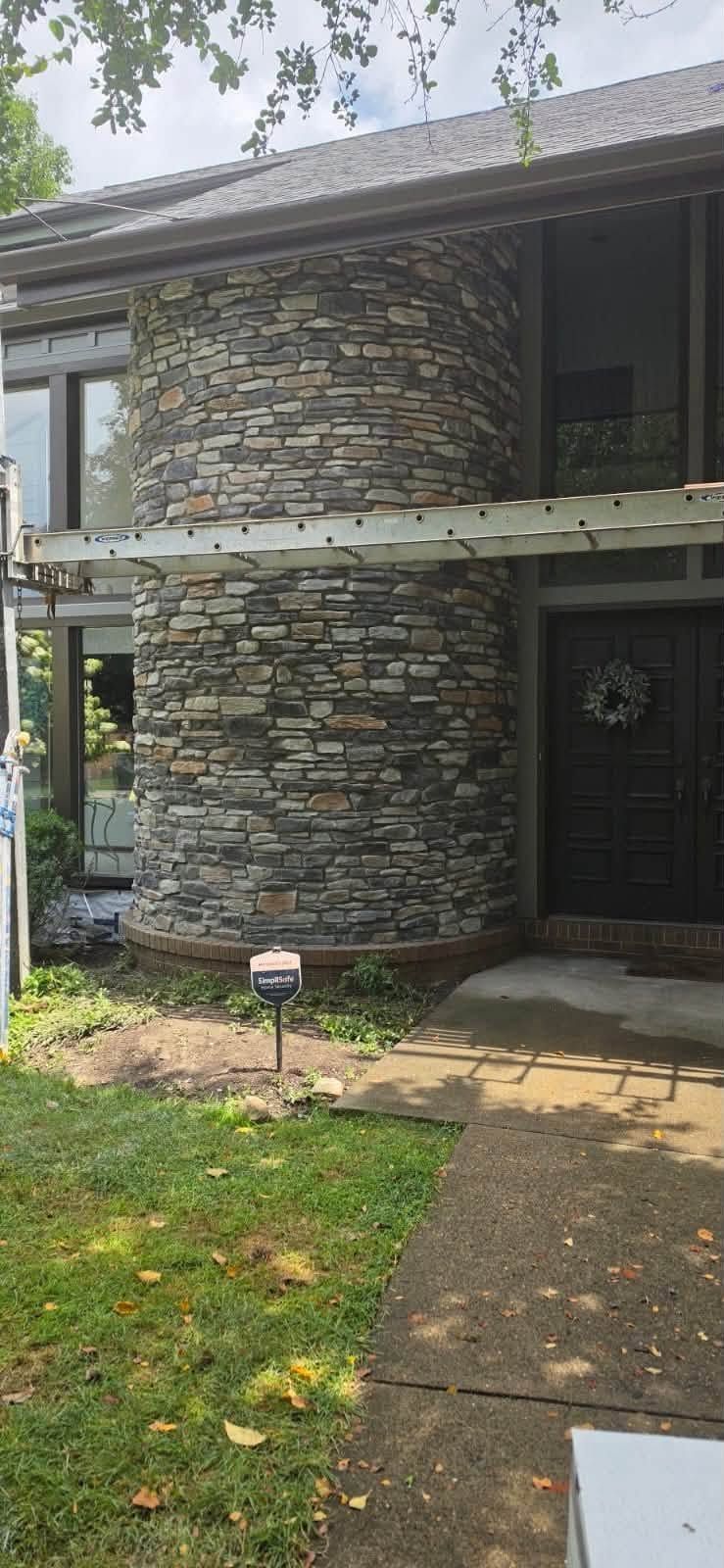 Front entrance of a stone-faced house with a black door, porch, and walkway through a grassy yard.