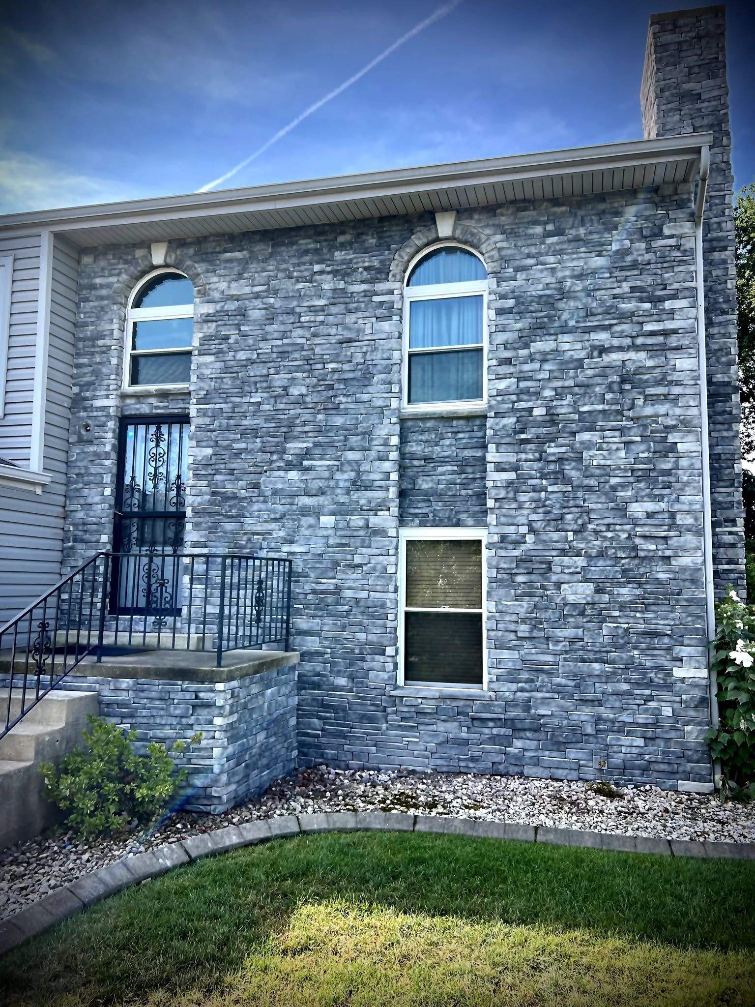 Stone house exterior with arched windows, front steps, and a small lawn under a blue sky