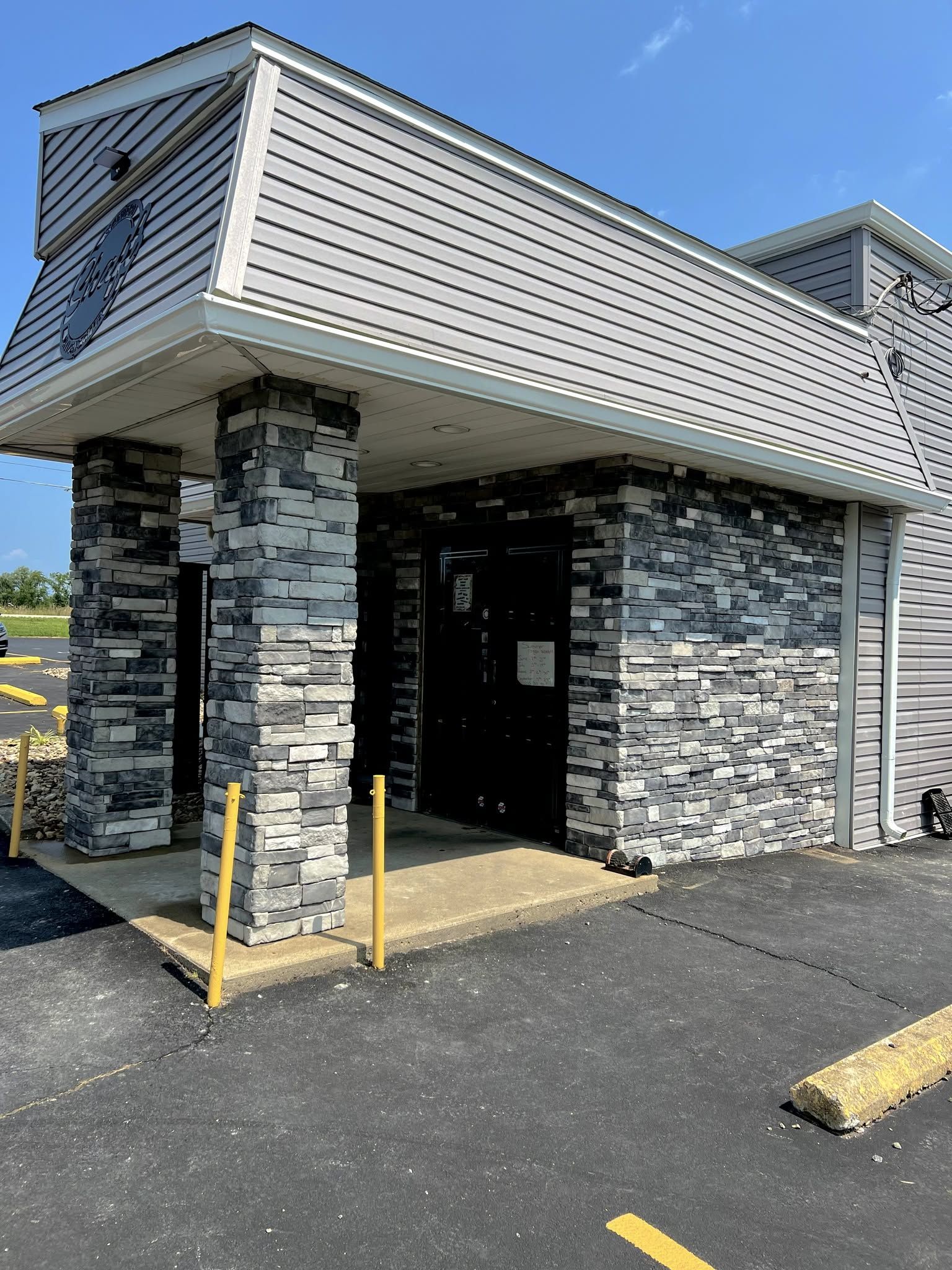 Covered building entrance with stone columns, gray siding, and yellow bollards beside an asphalt lot