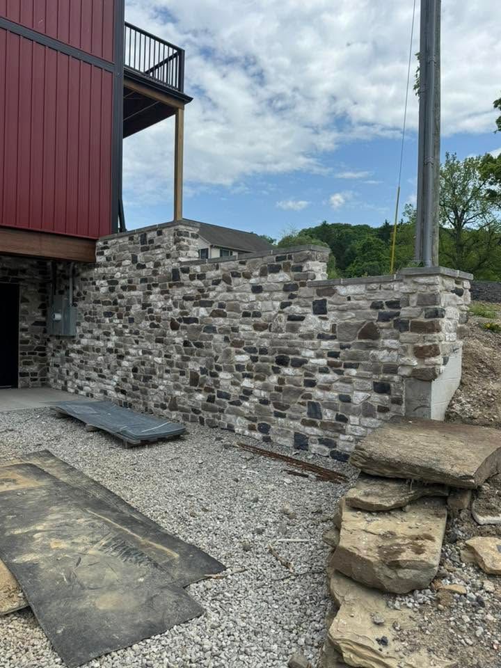 Stone retaining wall beside a red building with a balcony, gravel ground, and a cloudy sky.