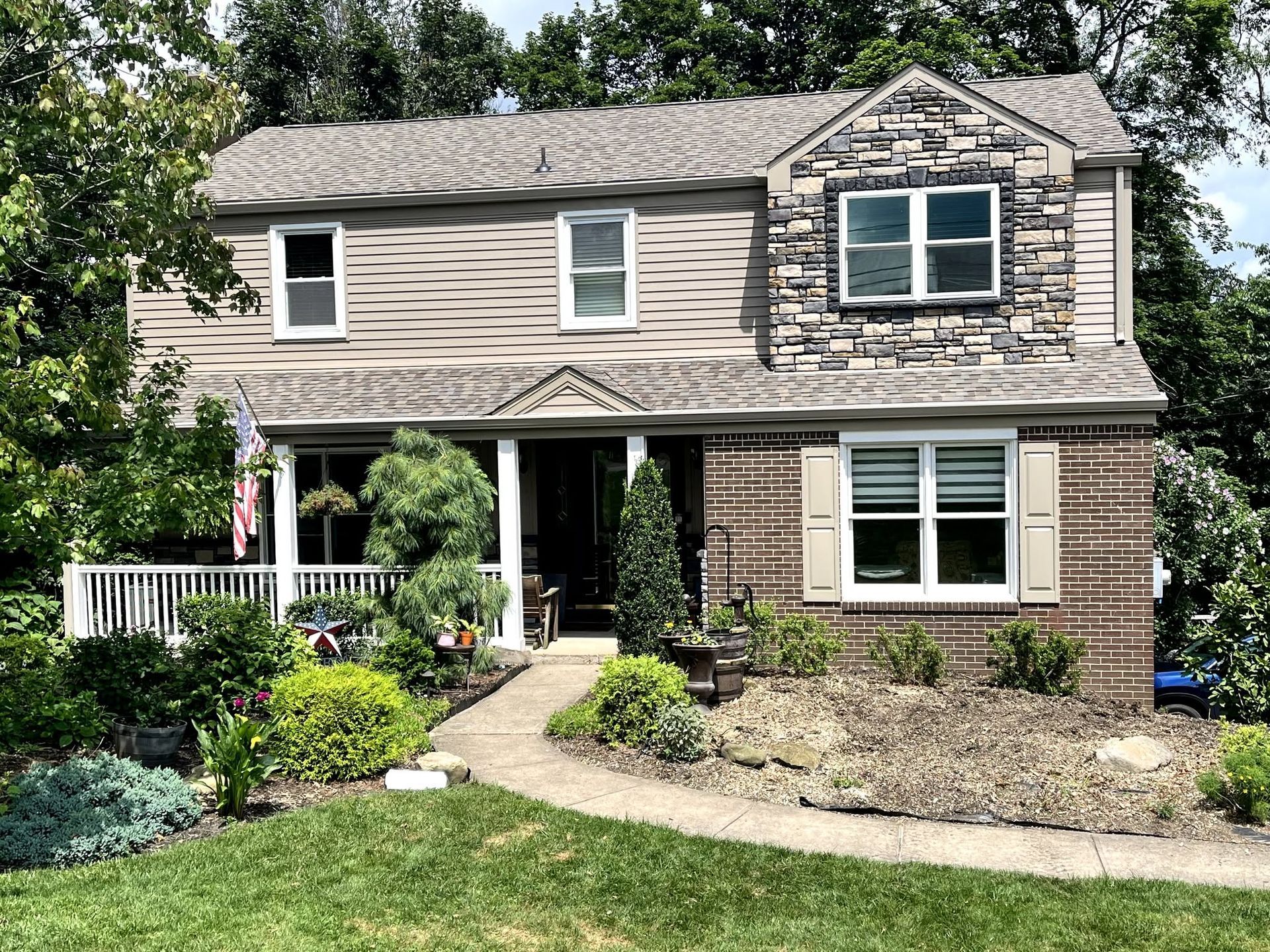 Two-story beige house with stone facade, front porch, and landscaped garden under trees