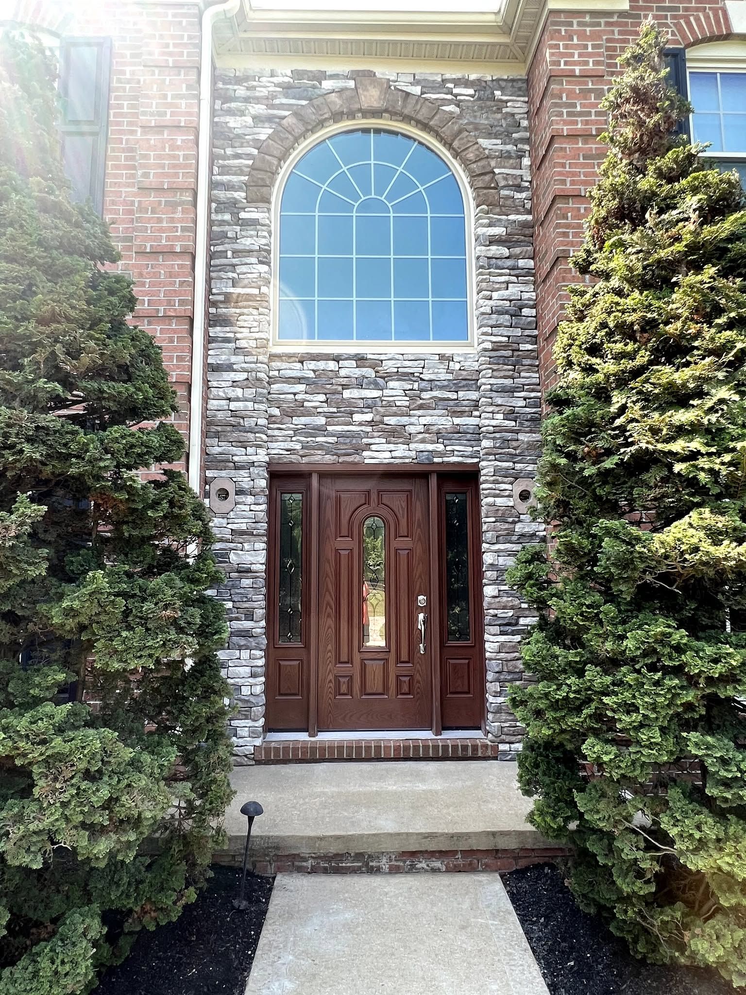 Front entrance with double wooden doors, arched window above, stone facade, and two tall evergreen shrubs.