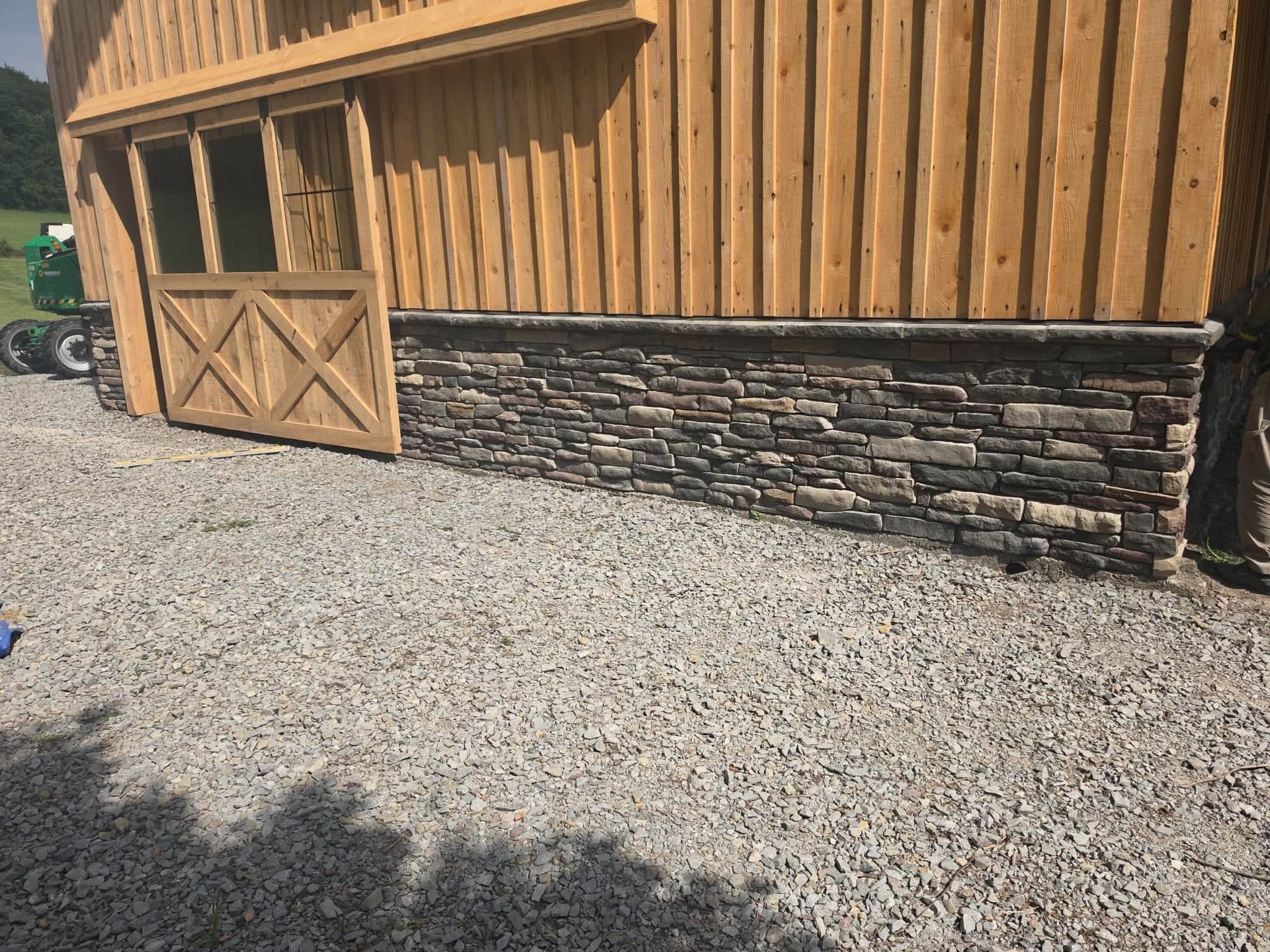 Wooden barn wall with stone base and open door beside a gravel yard, partly shaded by trees