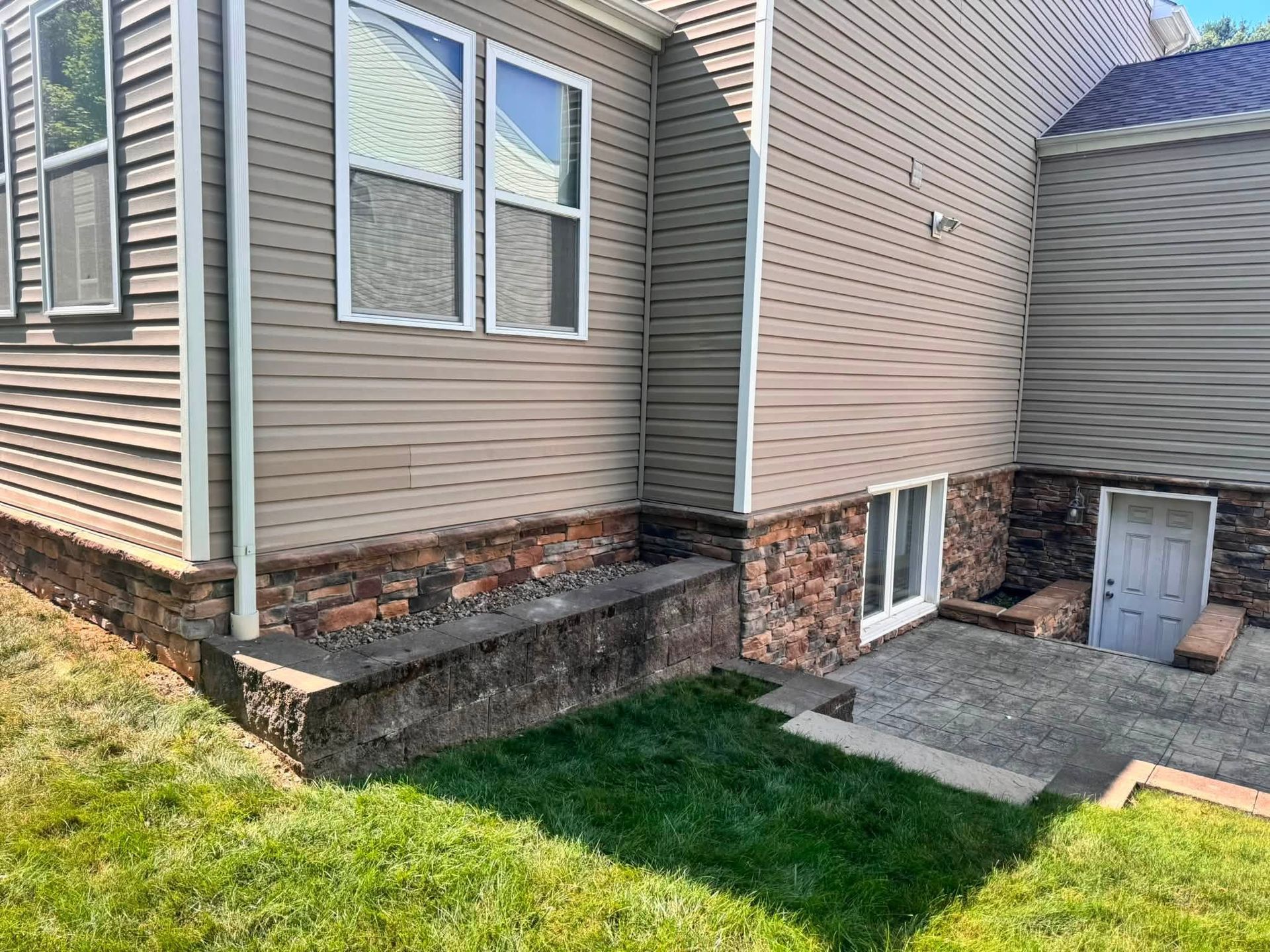 Residential home exterior with vinyl siding, basement windows, and a small concrete patio beside a grassy yard