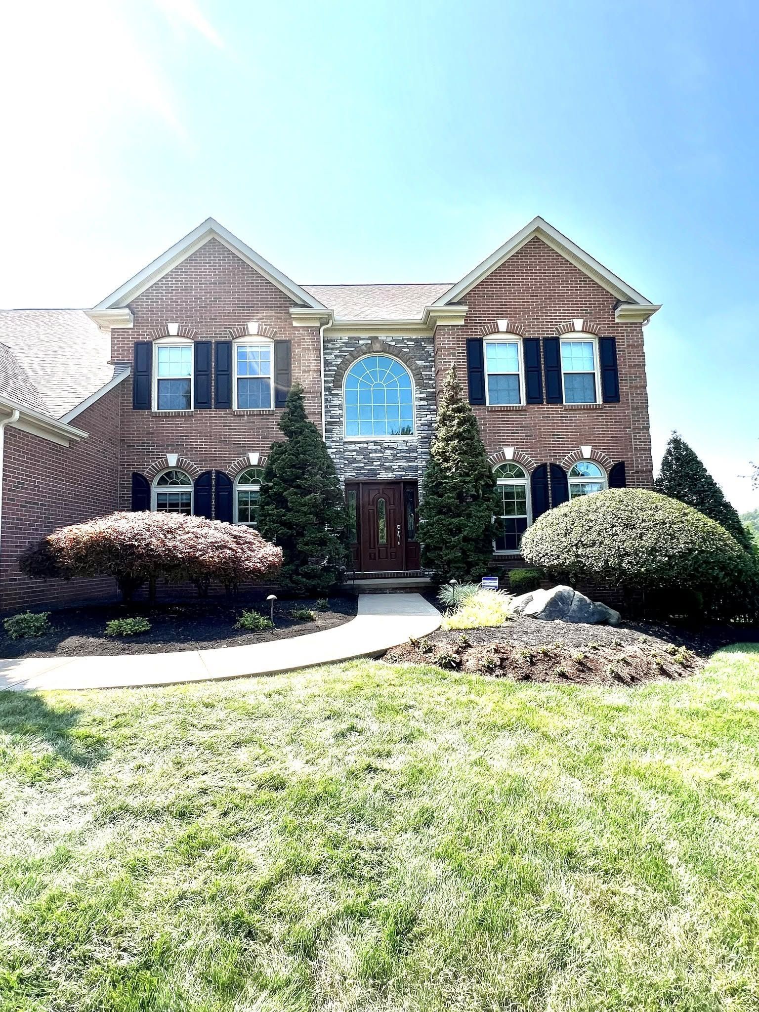 Brick two-story suburban house with peaked roofs, front door, windows, and landscaped shrubs in bright sunlight