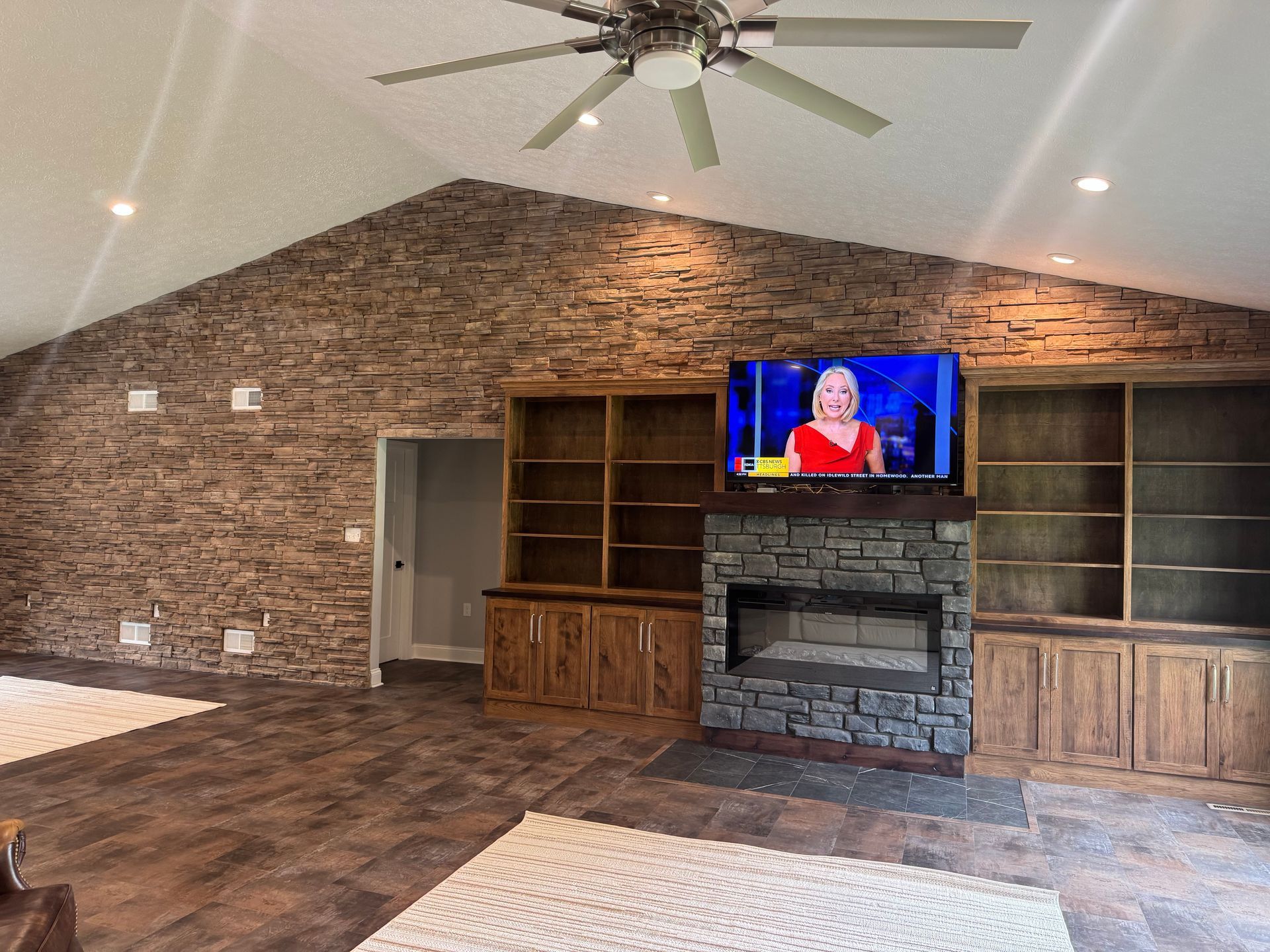 Spacious living room with stone fireplace, built-in cabinets, wood floor, and wall-mounted TV under a vaulted ceiling