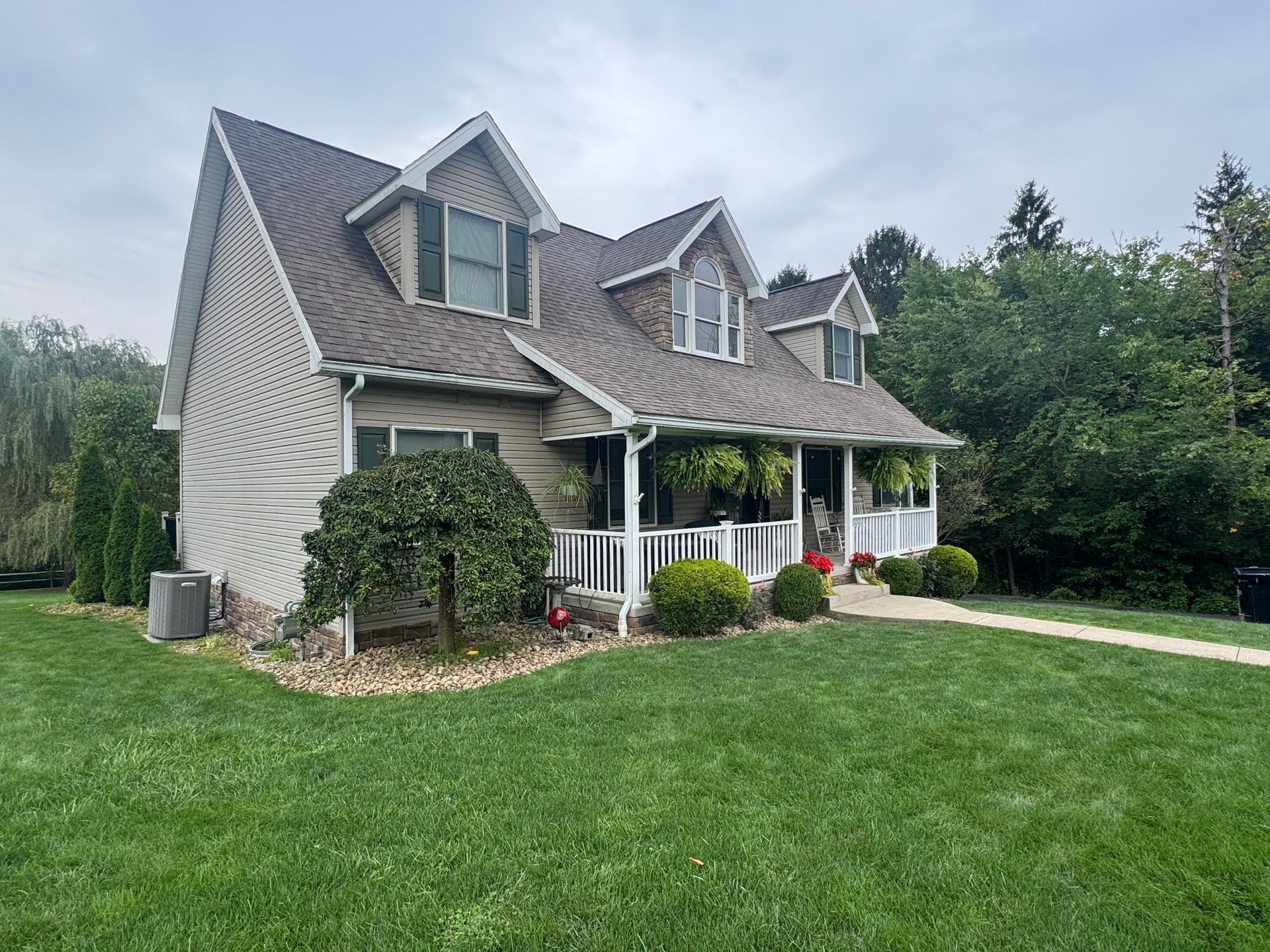 Gray house with white porch and dormer windows on a green lawn, surrounded by trees